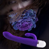 A woman with closed eyes delicately holds a vibrant purple flower near her face. In the foreground, the VeDO Wink Rabbit Vibe in Indigo, boasting a sleek, curved design and a white base, complements the serene essence of nature.